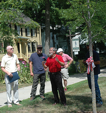 Bud Winsor (center, in red shirt) at the dedication of the Kentucky coffee tree (with red and white ribbon) to honor retiring Keene State arborist Jeff Garland (left, in creme shirt)