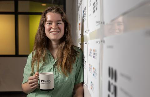 A female student, Julia Baer, stands in front of a white board holding a mug with the Cohen Institute for Holocaust & Genocide Studies logo on it