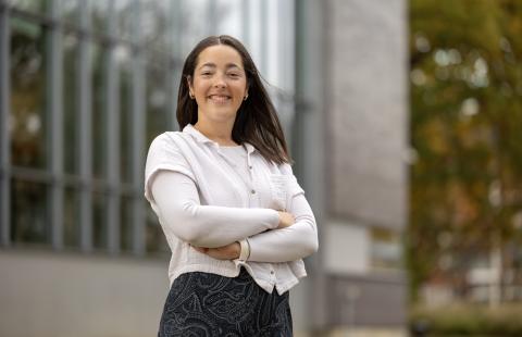 A woman with brunette hair and wearing a white blouse stands, arms crossed, smiling at the camera