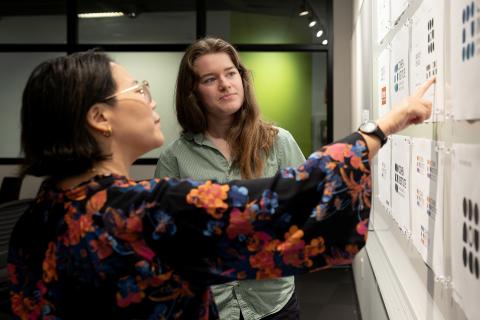 Two people look at paper pinned to a whiteboard